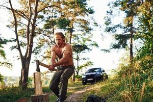 lenhador com um machado cortando madeira. homem bonito sem camisa com tipo de corpo musculoso está na floresta durante o dia foto