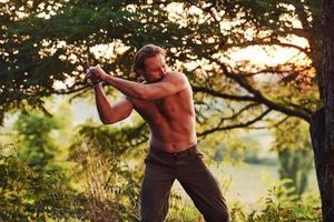 processo de cortar a árvore usando o machado. homem bonito sem camisa com tipo de corpo musculoso está na floresta durante o dia foto