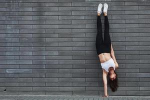 fazendo pino. jovem morena esportiva com corpo esbelto contra a parede de tijolos da cidade durante o dia foto