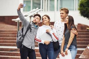 de pé e tirando selfie. grupo de jovens estudantes em roupas casuais perto da universidade durante o dia foto