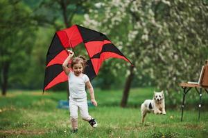 com cachorro fofo. criança do sexo feminino positiva correndo com pipa colorida vermelha e preta nas mãos ao ar livre foto