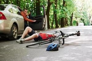 motorista abrindo a porta. vítima no asfalto. bicicleta e acidente de carro prateado na estrada na floresta durante o dia