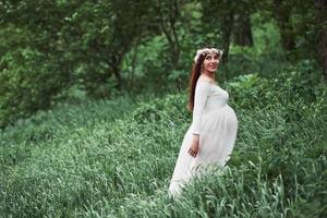 na floresta. linda mulher grávida de vestido dar um passeio ao ar livre. morena positiva foto