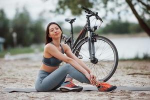 relaxado e satisfeito. ciclista feminina com boa forma corporal sentada perto de sua bicicleta na praia durante o dia foto