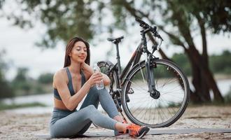 água potável. ciclista feminina com boa forma corporal sentada perto de sua bicicleta na praia durante o dia