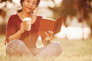 garota amigável satisfeita com o clima, café e livro novo. alegre mulher afro-americana no parque no verão foto