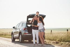 garoto senta-se nos ombros do pai. a família se diverte no campo perto do automóvel prateado ao pôr do sol foto