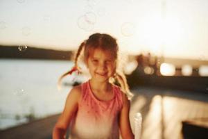 sol incrível. menina feliz brincando com bolhas perto do lago no parque foto