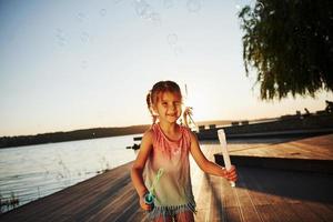 tempo da noite. menina feliz brincando com bolhas perto do lago no parque foto