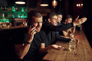 homem fala pelo telefone. três fãs de esportes em um bar assistindo futebol. com cerveja nas mãos foto