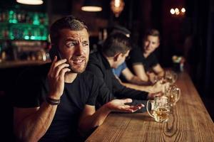 homem fala pelo telefone. três fãs de esportes em um bar assistindo futebol. com cerveja nas mãos foto