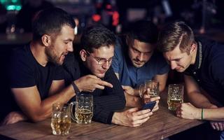 conteúdo engraçado no smartphone. três amigos descansando no bar com cerveja nas mãos. conversando foto