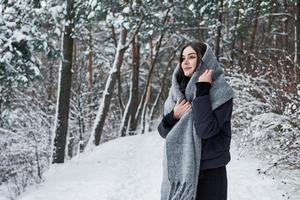 aquecendo. retrato de mulher encantadora na jaqueta preta e cachecol cinza na floresta de inverno foto