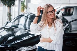 suas chaves. jovem gerente na loja de automóveis se posiciona contra um carro preto perfeitamente polido foto