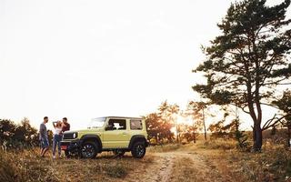 cena rural. grupo de amigos alegres tem um bom fim de semana em um dia ensolarado perto de seu carro verde ao ar livre foto