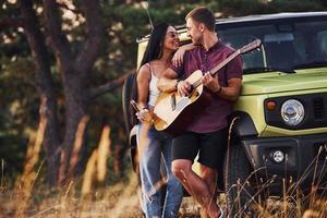 casal tem fim de semana ao ar livre em dia ensolarado. pessoas adoráveis ficam perto de jipe verde com álcool e violão foto