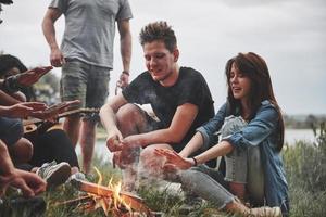 sentado perto do fogo. grupo de pessoas faz piquenique na praia. amigos se divertem no fim de semana foto