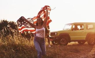 garota corre para frente. amigos tenham um bom fim de semana ao ar livre perto do carro verde deles com a bandeira dos eua foto