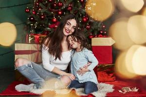 eles sentem a satisfação de estarem juntos nessas férias. alegre mãe e filha sentada perto da árvore de natal que atrás. retrato bonito foto