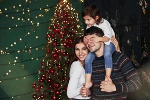 mamãe está sorrindo. menina senta-se nos ombros do pai e cobre os olhos. natal decorado ao fundo foto