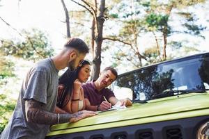 jovens amigos lendo o mapa que está no capô do jipe verde moderno na floresta foto