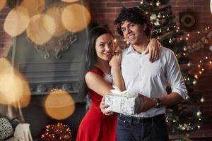 Este presente é para você. lindo casal comemorando o ano novo na sala decorada com árvore de natal e lareira atrás foto
