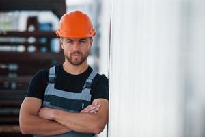 encostado na parede. trabalhador industrial sério dentro de casa na fábrica. jovem técnico com capacete laranja foto