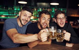 copos batendo. três fãs de esportes em um bar assistindo futebol. com cerveja nas mãos