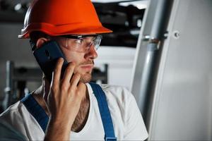 usando telefone. retrato de engenheiro em fábrica metalúrgica em óculos e capacete de proteção foto