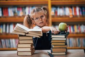 menina bonitinha com tranças está na biblioteca. maçã nos livros foto