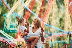 tomando sorvete. menina alegre sua mãe se diverte no parque juntos perto de atrações foto