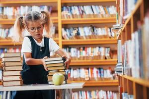 menina bonitinha com tranças está na biblioteca. maçã nos livros foto