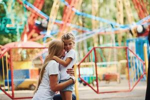 abraçando um ao outro. menina alegre sua mãe se diverte no parque juntos perto de atrações foto