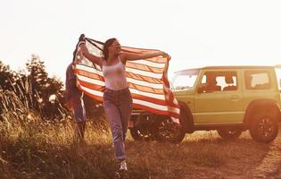 garota corre para frente. amigos tenham um bom fim de semana ao ar livre perto do carro verde deles com a bandeira dos eua foto