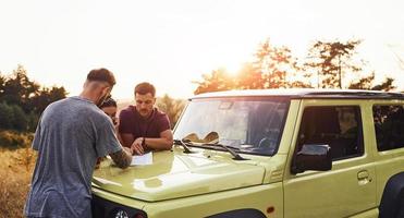 gente lendo mapa que jaz no capô do automóvel. grupo de amigos alegres tem um bom fim de semana em um dia ensolarado perto de seu carro verde ao ar livre foto