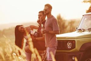 tocando violão e cantando. grupo de amigos alegres tem um bom fim de semana em um dia ensolarado perto de seu carro verde ao ar livre foto