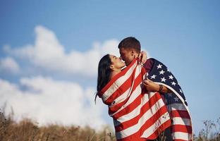 beijando um ao outro. sente liberdade. lindo casal com bandeira americana se diverte ao ar livre no campo foto