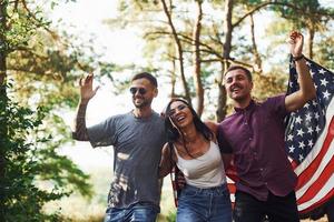 na floresta. amigos tenham um bom fim de semana ao ar livre perto do carro verde deles com a bandeira dos eua foto