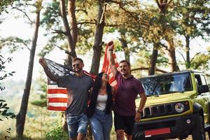 na floresta. amigos tenham um bom fim de semana ao ar livre perto do carro verde deles com a bandeira dos eua foto