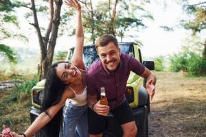 sorrindo e se divertindo. casal de jovens com álcool se divertem na floresta. jipe verde atrás foto