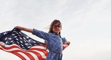 criança feminina patriótica com bandeira americana nas mãos. contra o céu nublado foto