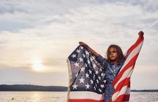 criança feminina patriótica com bandeira americana nas mãos. contra o céu nublado foto
