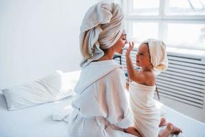 usando creme para limpar a pele. jovem mãe com sua filha tem dia de beleza dentro de casa no quarto branco foto