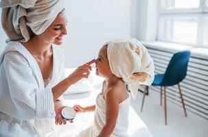 usando creme para limpar a pele. jovem mãe com sua filha tem dia de beleza dentro de casa no quarto branco foto