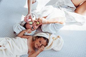 jovem mãe positiva e filha com toalha na cabeça deitada na cama junto com pepino foto