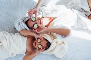 jovem mãe positiva e filha com toalha na cabeça deitada na cama junto com pepino foto