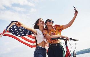 duas mulheres alegres patrióticas com bicicleta e bandeira dos eua nas mãos fazem selfie foto