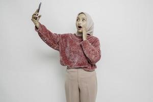 mulher asiática chocada vestindo suéter rosa e hijab, segurando seu telefone, isolado por fundo branco foto