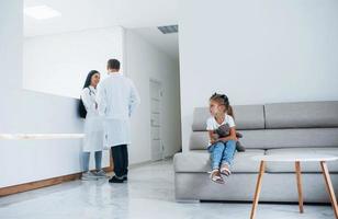 médicos atrás. menina bonitinha com ursinho de pelúcia nas mãos senta-se na sala de espera do hospital foto