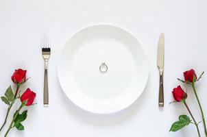 anel de diamante coloca no prato com faca, garfo e rosas vermelhas sobre fundo branco para o conceito de dia dos namorados. foto